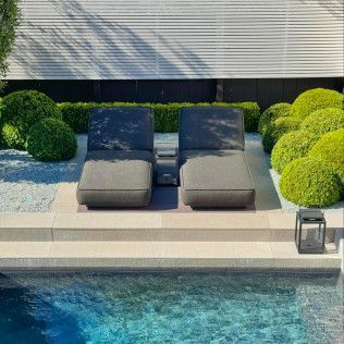 Two gray lounge chairs by a pool, with green shrubs and a white wall in the background.