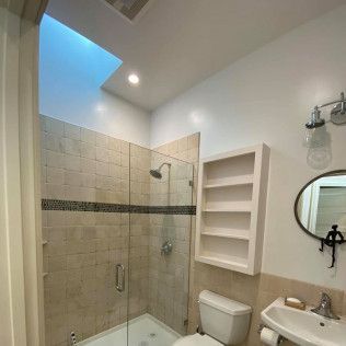 Bright bathroom with glass shower, white toilet, sink, wall shelves, and round mirror under a skylight.