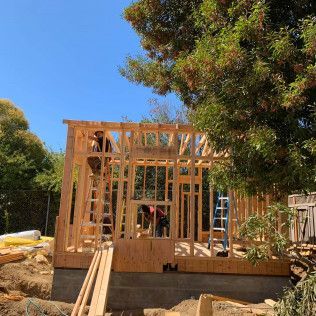 Wood-framed house under construction with workers, in a sunny yard with trees and blue sky