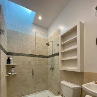 Small beige tiled bathroom with glass shower, skylight, toilet, sink, and wall shelves