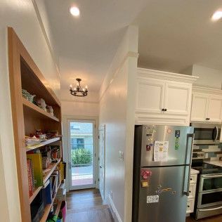 Narrow galley kitchen with white cabinets, stainless steel fridge and oven, and a bookshelf along the left wall.