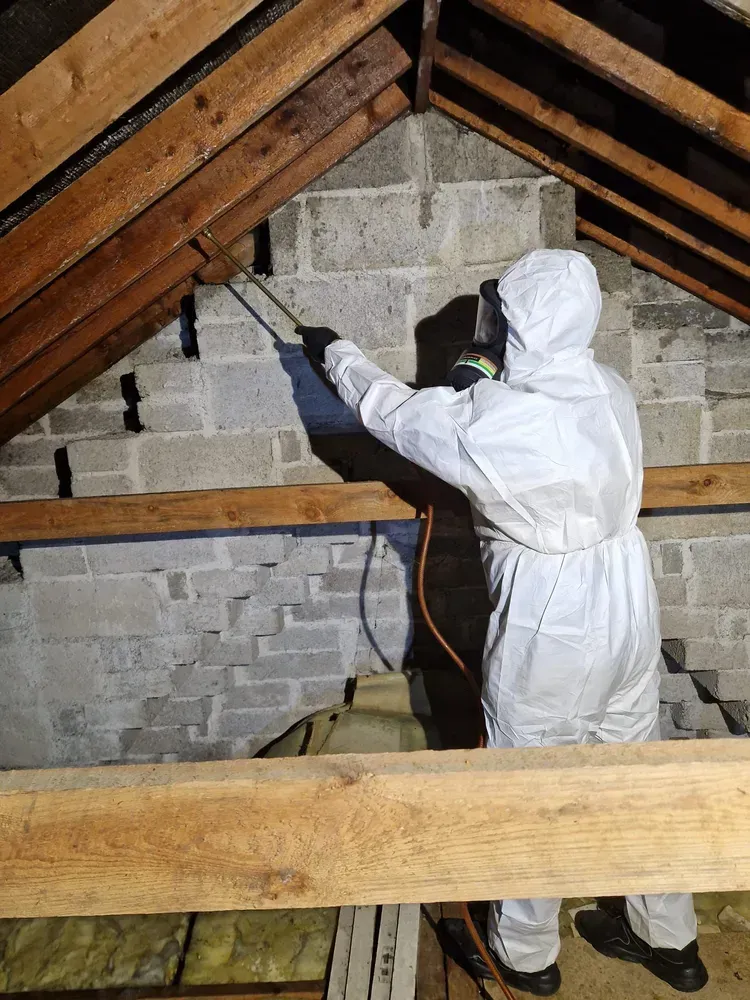 Person in white protective suit spraying insulation in an attic with exposed wooden beams and cinder block walls.