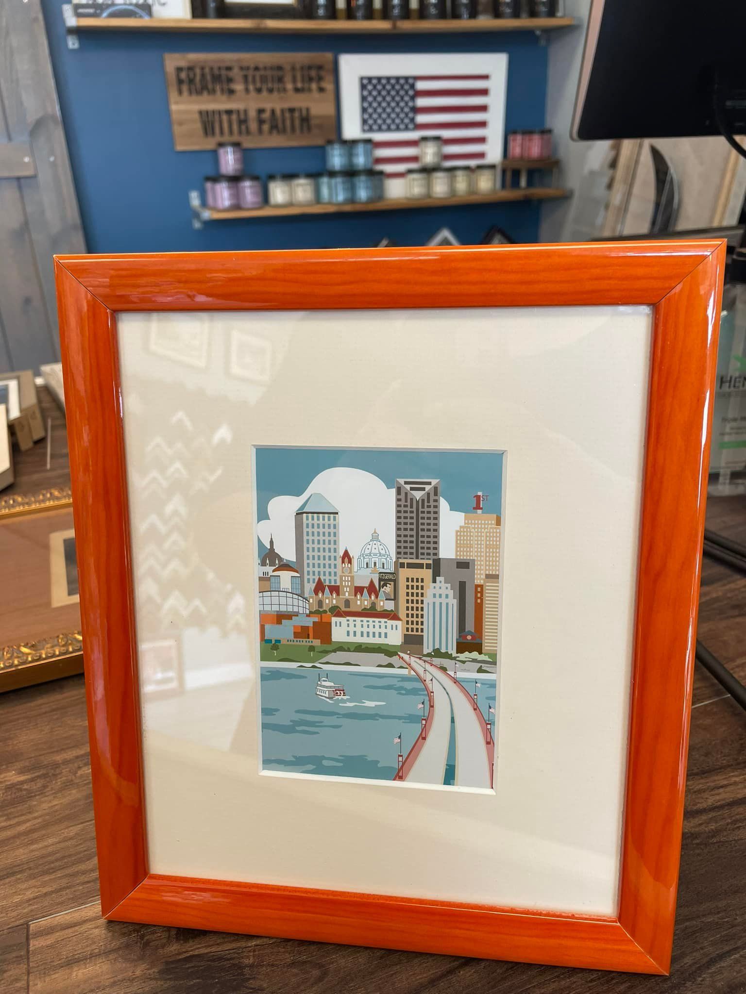 A framed illustration of a city skyline with a bridge and water, set on a wooden table in a shop.