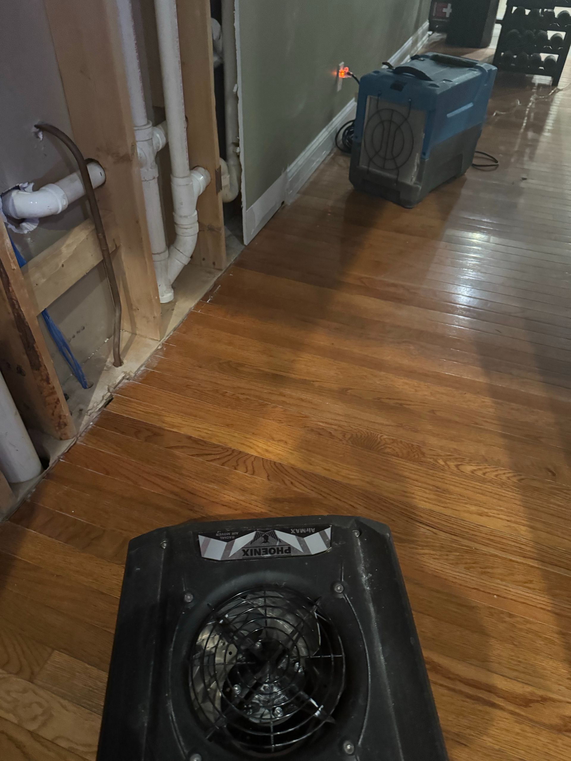 Damaged hallway floor with a black box fan in the foreground and a blue machine near the wall.