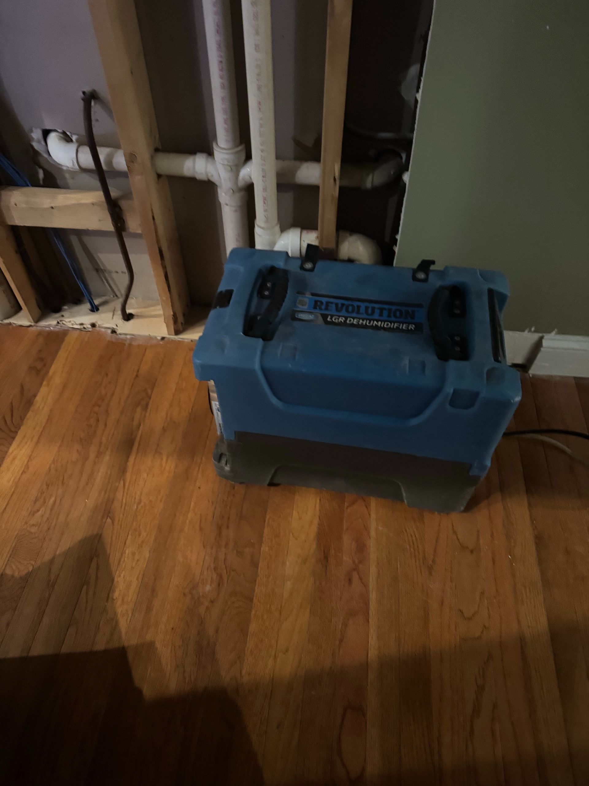 Blue toolbox on a hardwood floor beside exposed pipes near a wall opening.