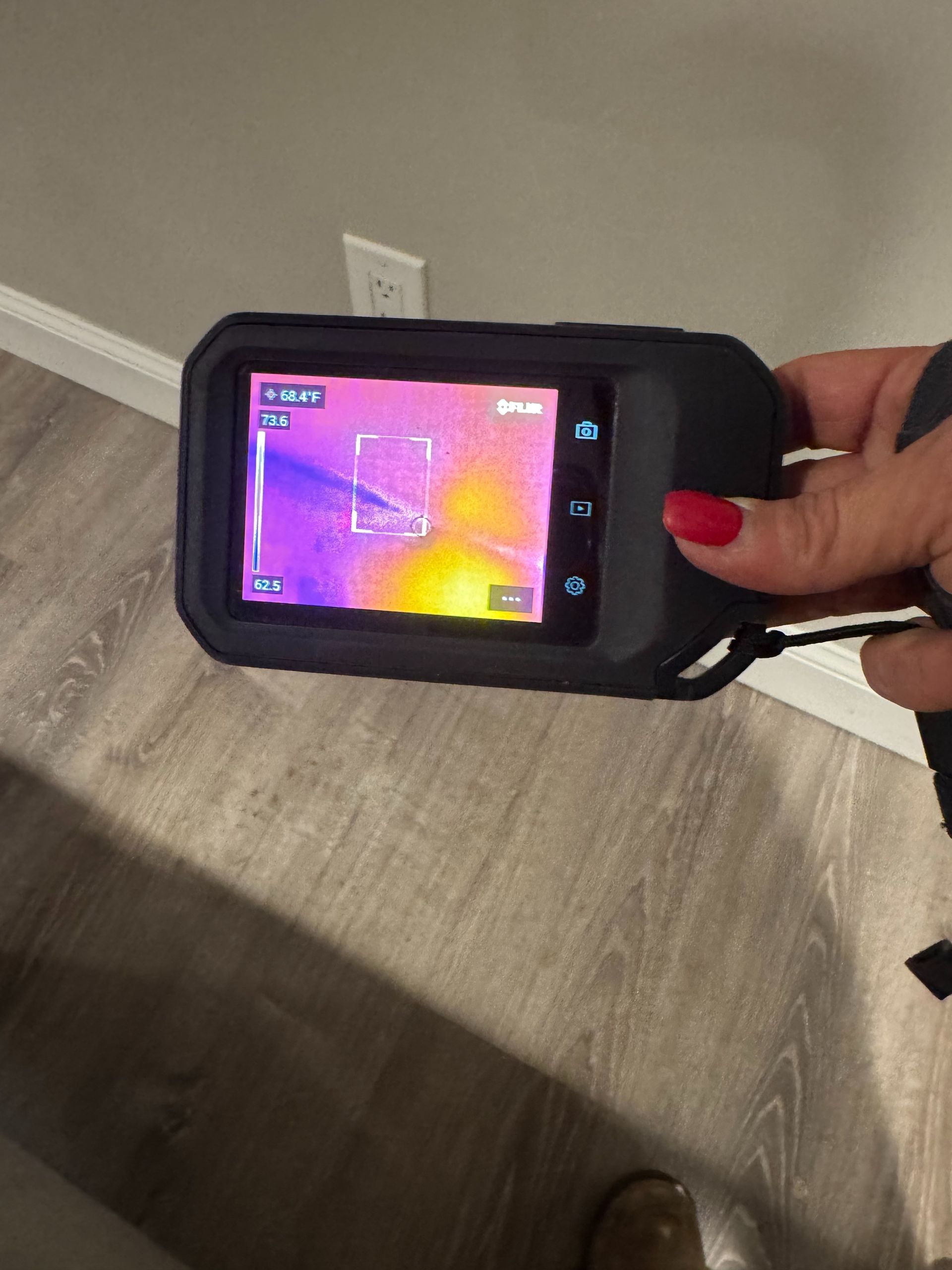 Handheld thermal camera showing a colorful heat map in front of a wall outlet on a wooden floor