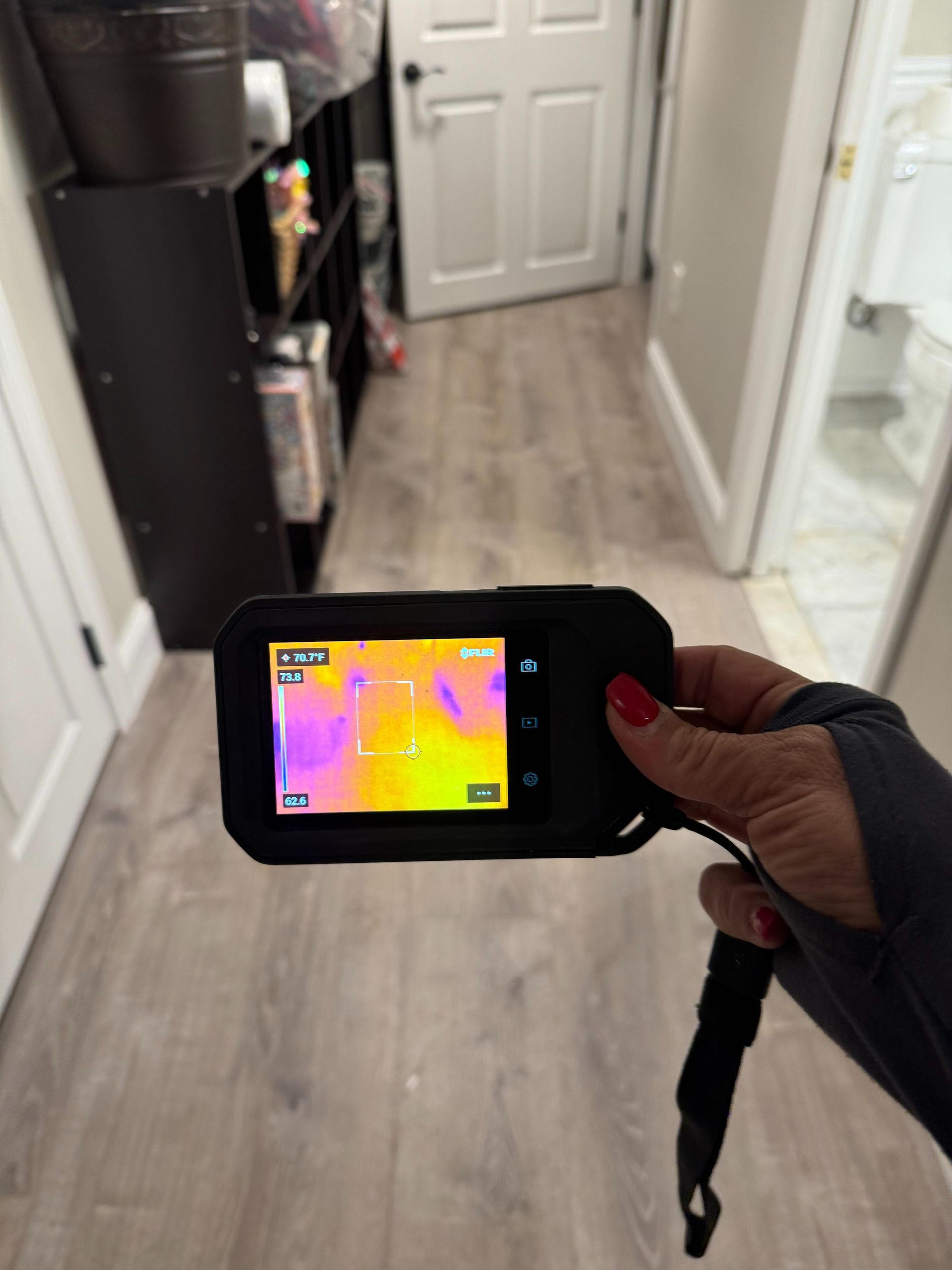 Hand holding a thermal camera showing a pink heat map in a hallway with a white door and glass shower.