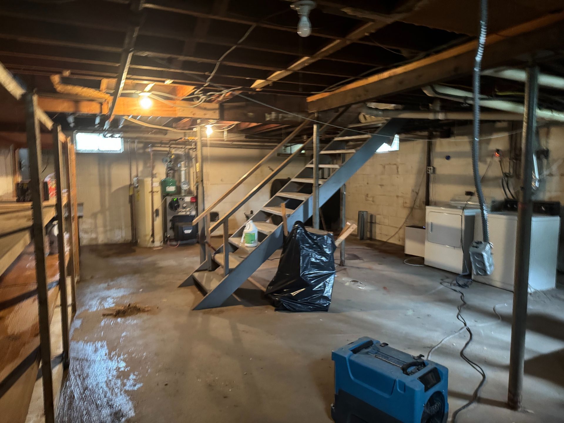 Dim basement with exposed pipes, a staircase, storage shelves, and a blue toolbox on the floor.