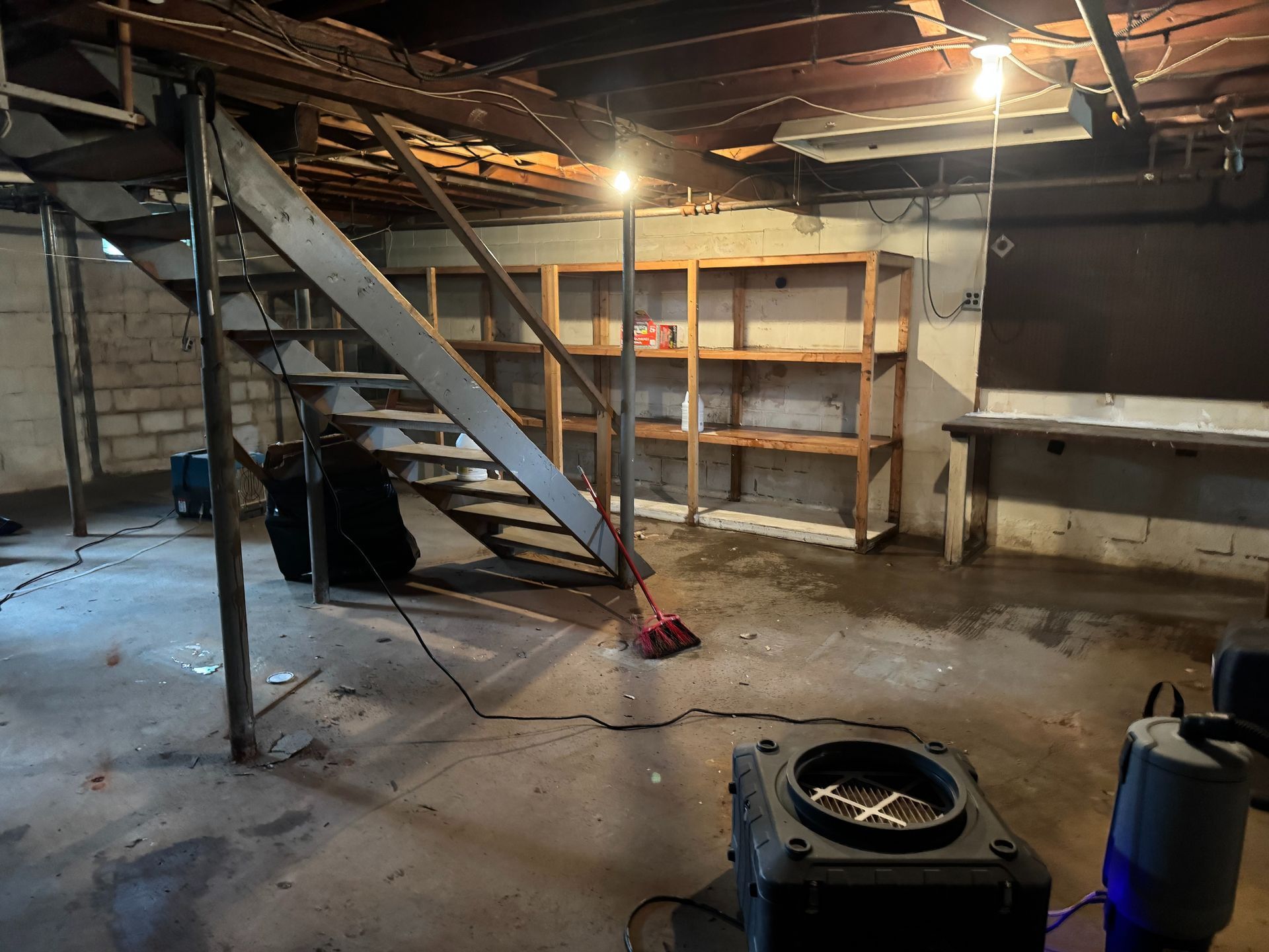 Dim basement with metal stairs, shelves, concrete floor, and cleaning supplies under warm overhead lights