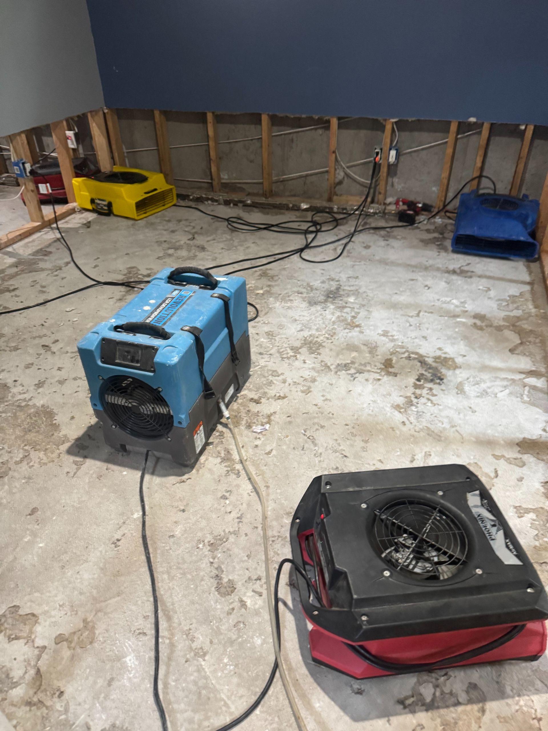 Workshop floor with two portable air movers, cords, and a yellow machine against exposed wall framing.