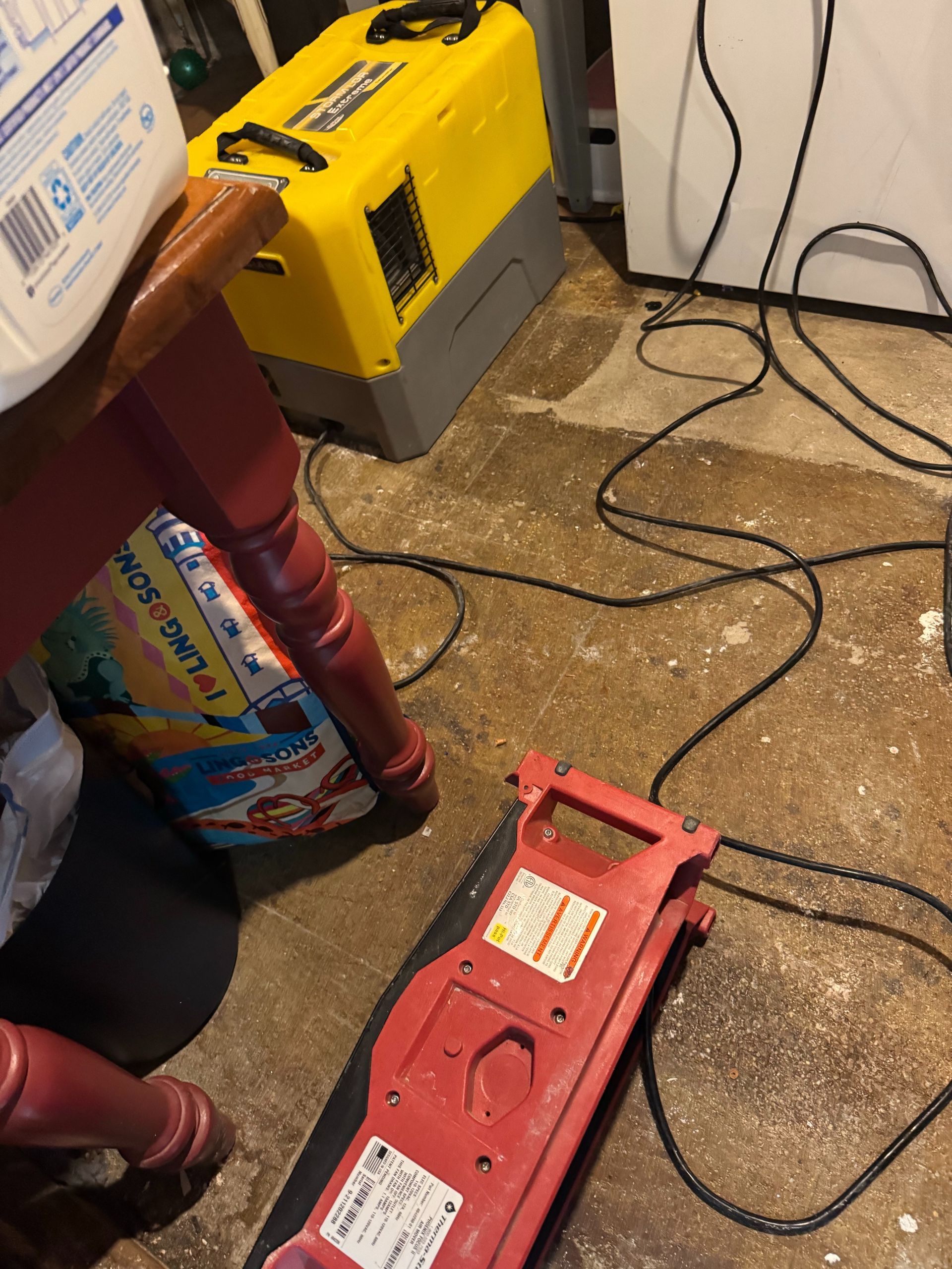 Red jump starter on a cluttered floor beside a yellow battery charger and tangled black cables
