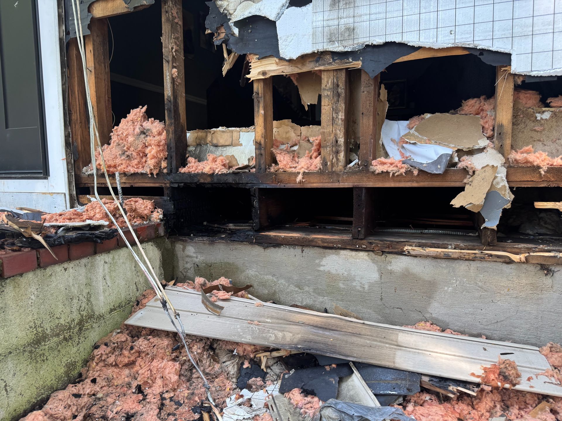 Damaged house exterior with collapsed wall, broken siding, and debris piled against the foundation.