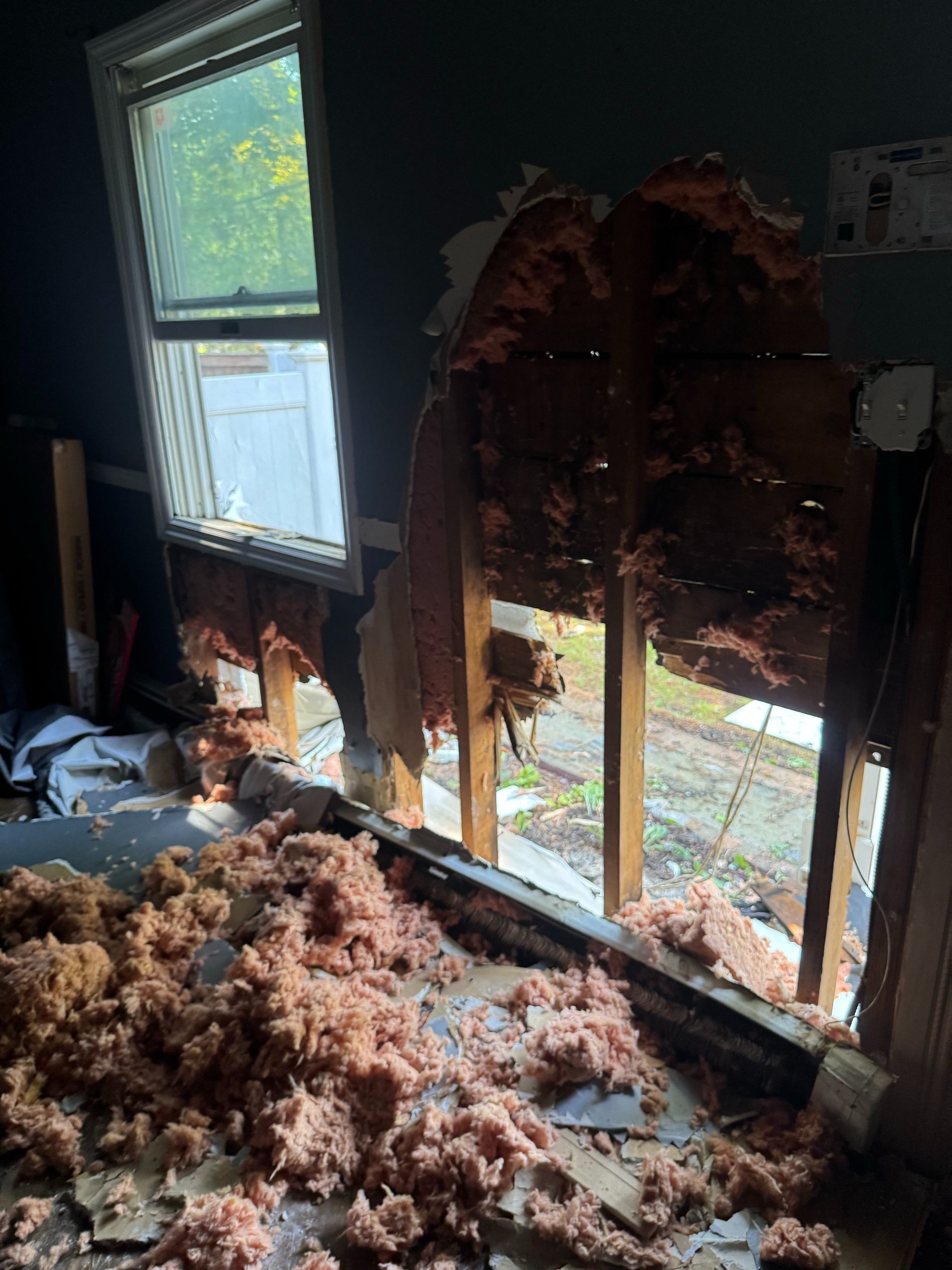 Damaged room with torn insulation, exposed wall studs, and broken windows letting in daylight.