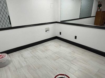 Empty tiled room corner with white walls, black trim, and a mirror along the upper wall.