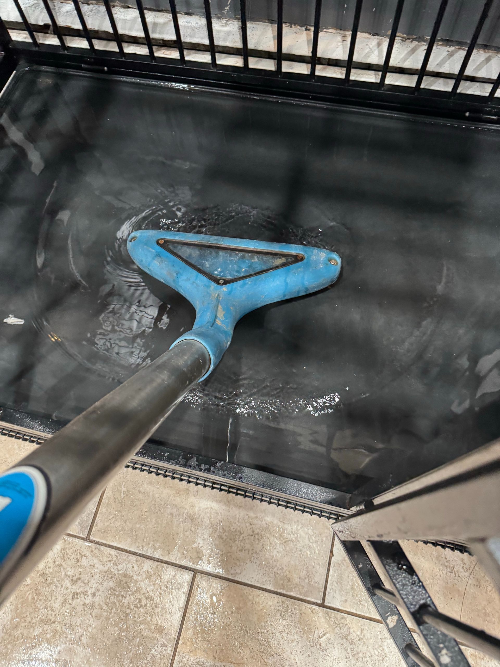 Blue floor-cleaning brush scrubbing a wet black tiled surface near a metal gate and tiled floor