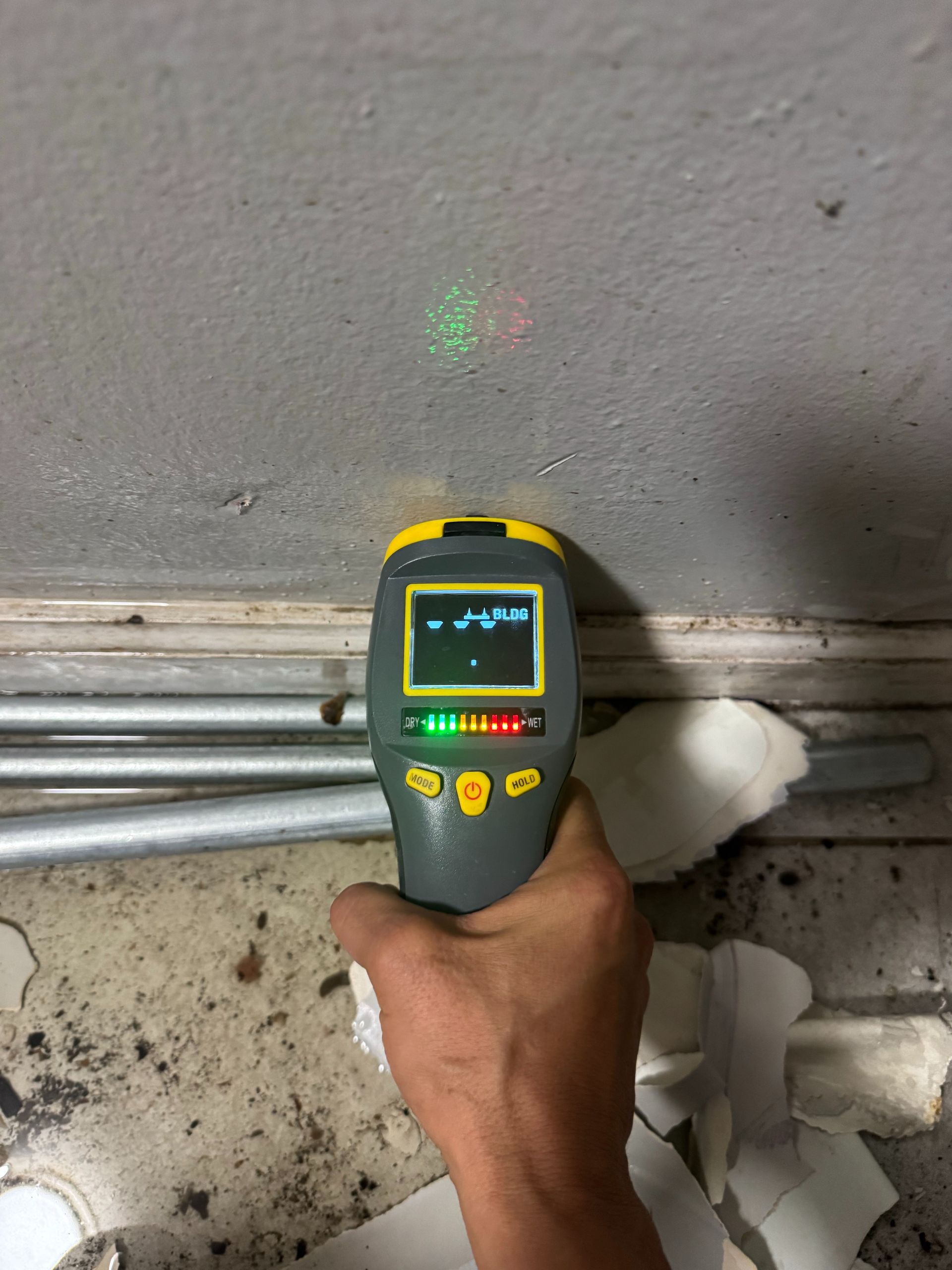 Handheld laser level measuring a gray wall above metal pipes and debris on a concrete floor