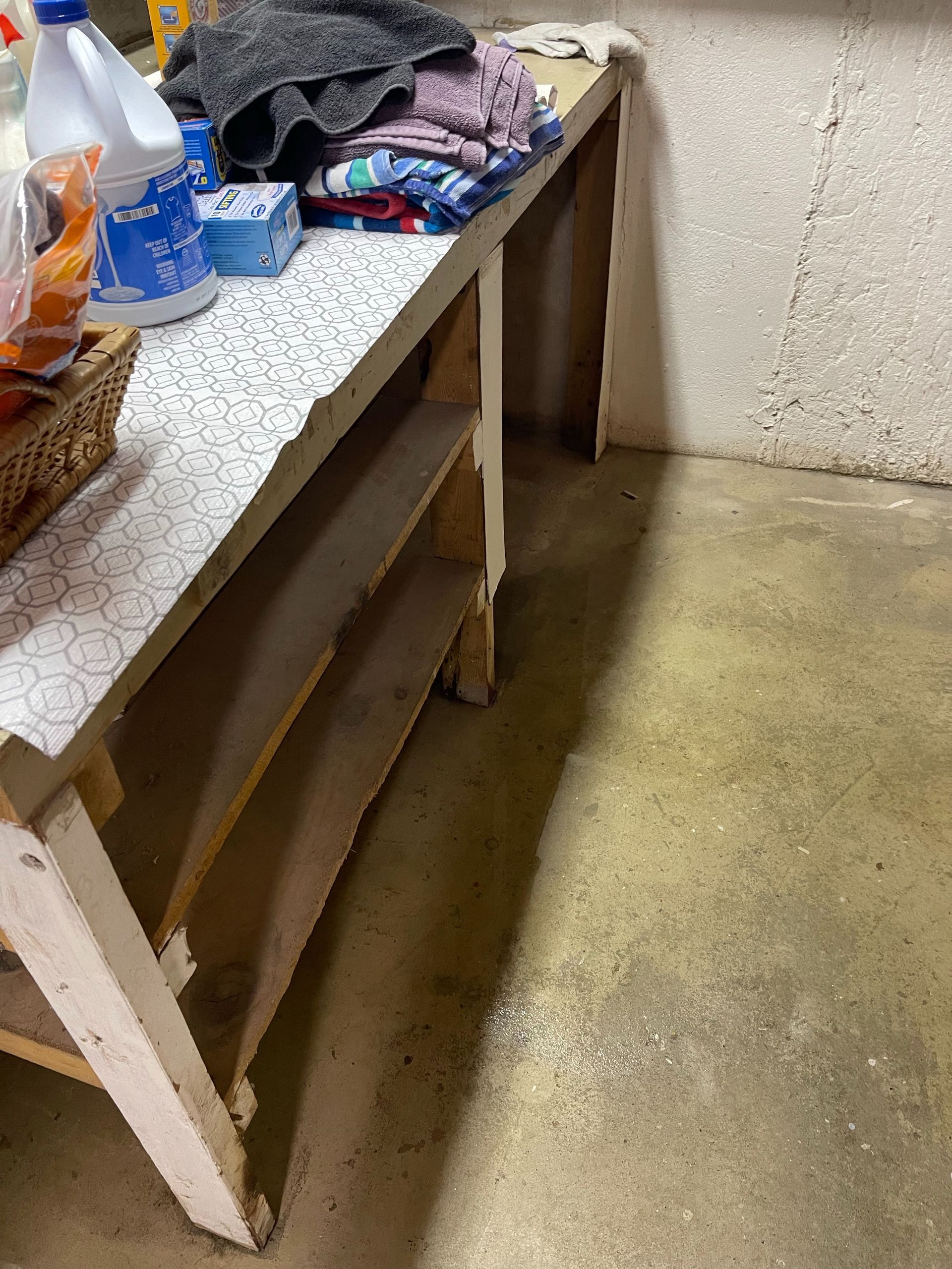 Cluttered workbench with bottles and folded clothes in a small room with a bare concrete floor.