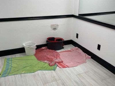 Cleaning supplies and towels on the floor in a room corner with a white bucket and black basin.