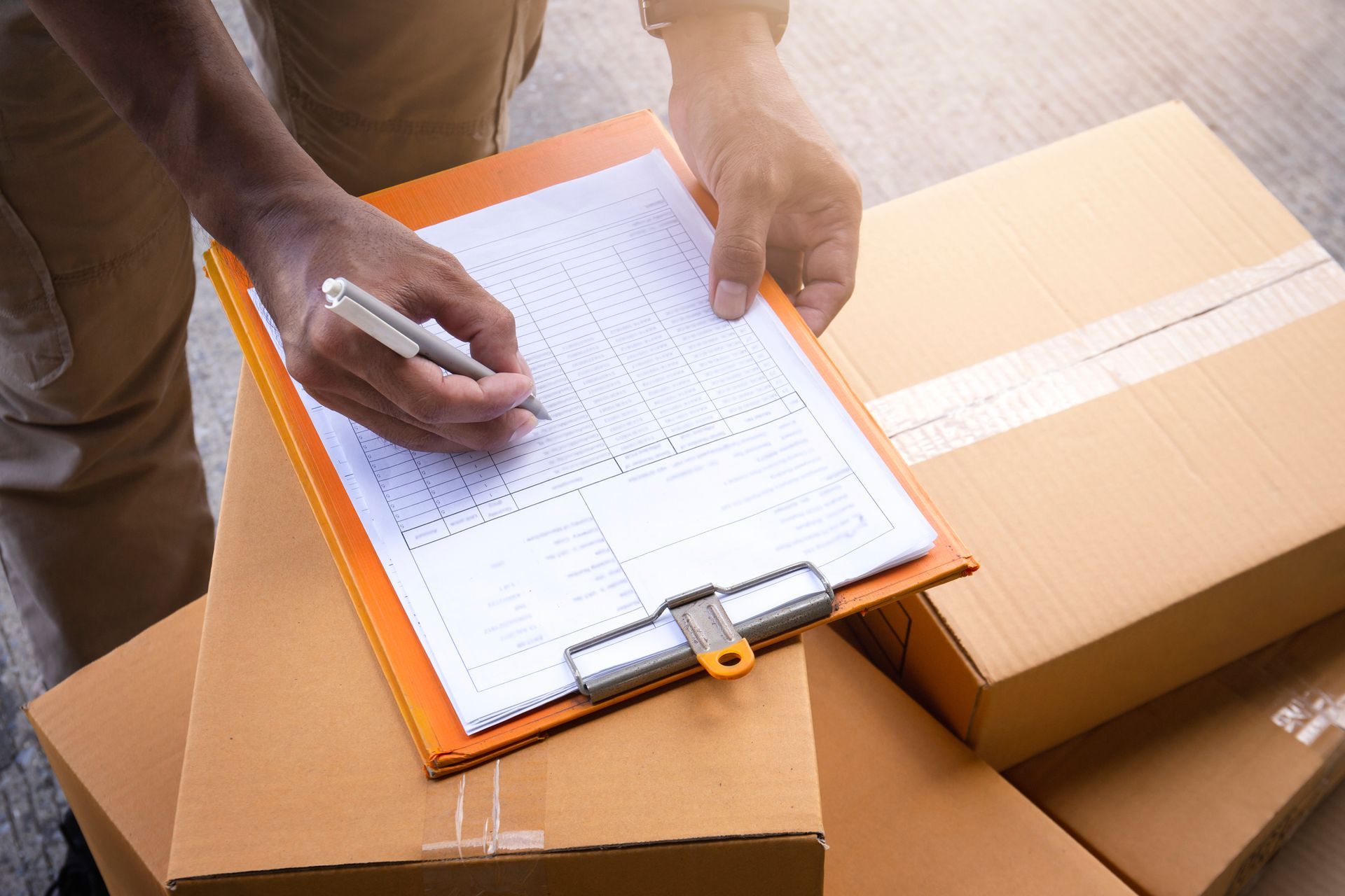 A person is writing on a clipboard next to a pile of cardboard boxes.