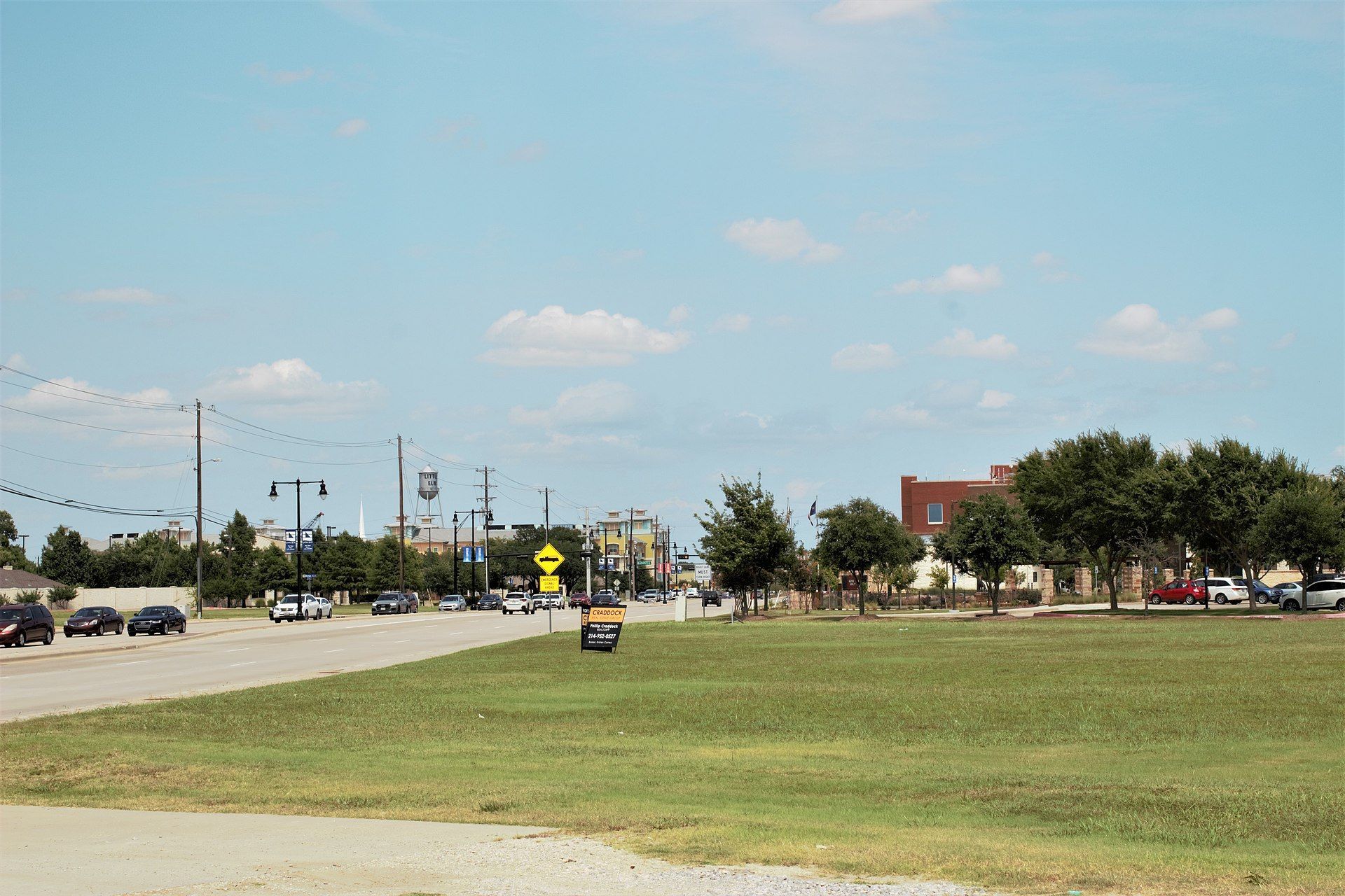 A grassy roadside beside a street with cars, trees, and a partly cloudy sky.