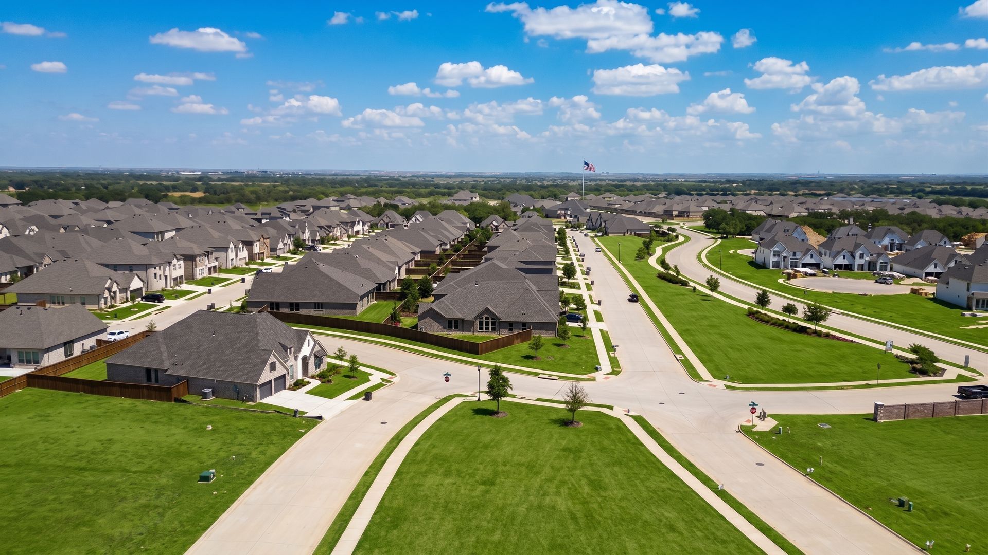 Aerial view of a suburban neighborhood with rows of houses, curving streets, and green lawns under a blue sky.