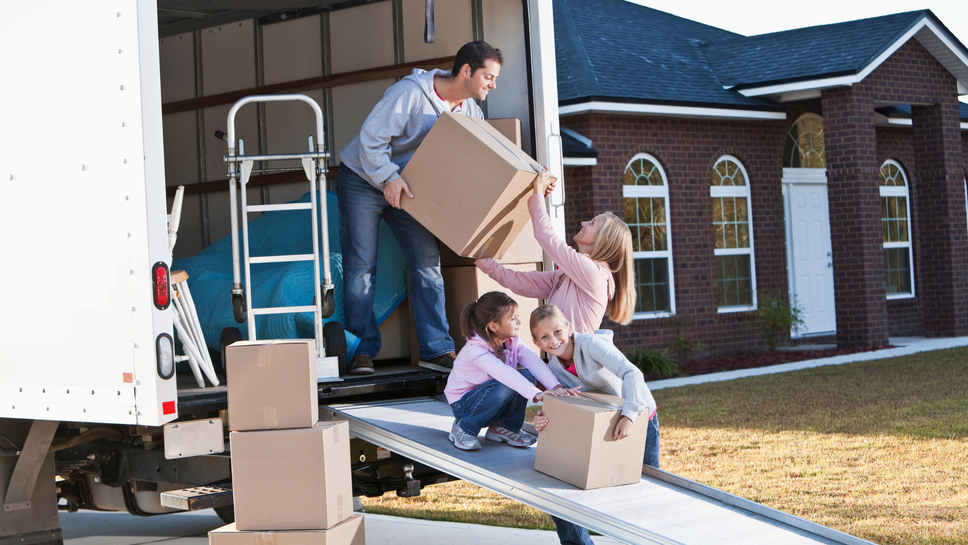 Mover unloading boxes from a truck onto a ramp with help from two children outside a house
