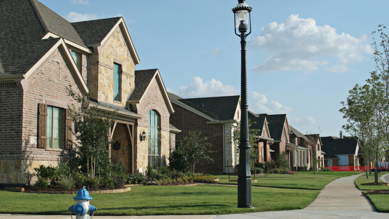 Suburban neighborhood street with brick houses, sidewalk, lamppost, and a blue fire hydrant under a clear sky