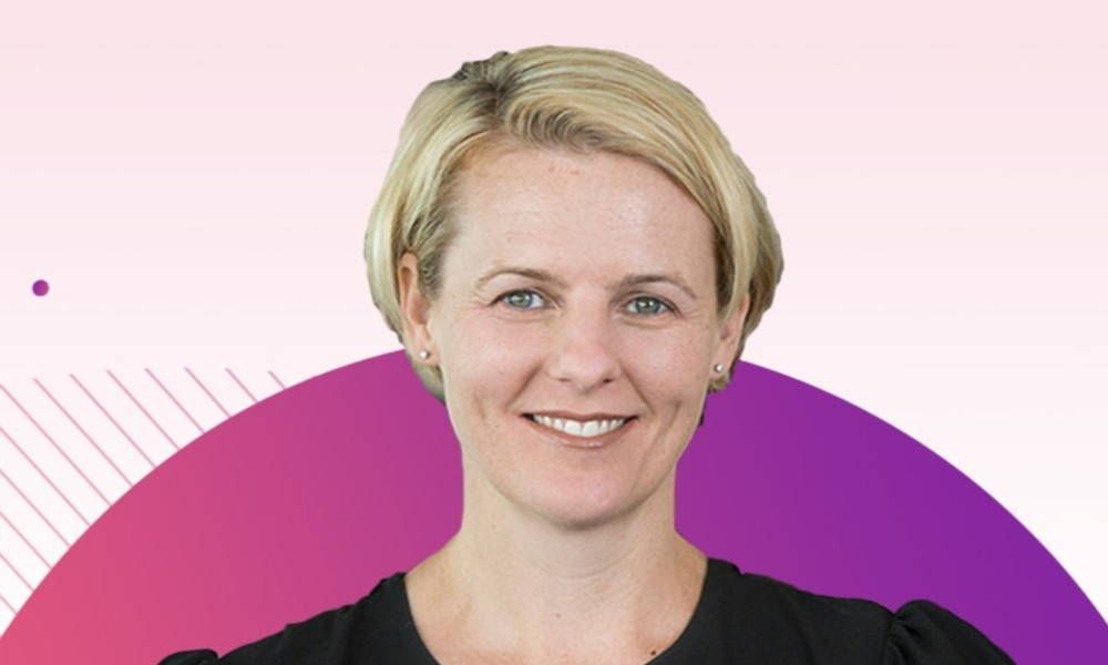 A woman is smiling in front of a pink and purple background.