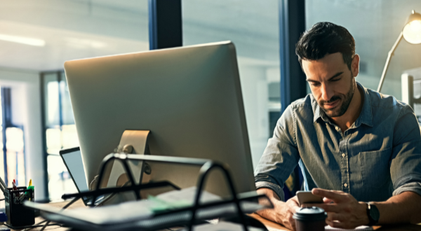 A man is sitting at a desk in front of a computer and looking at his phone.