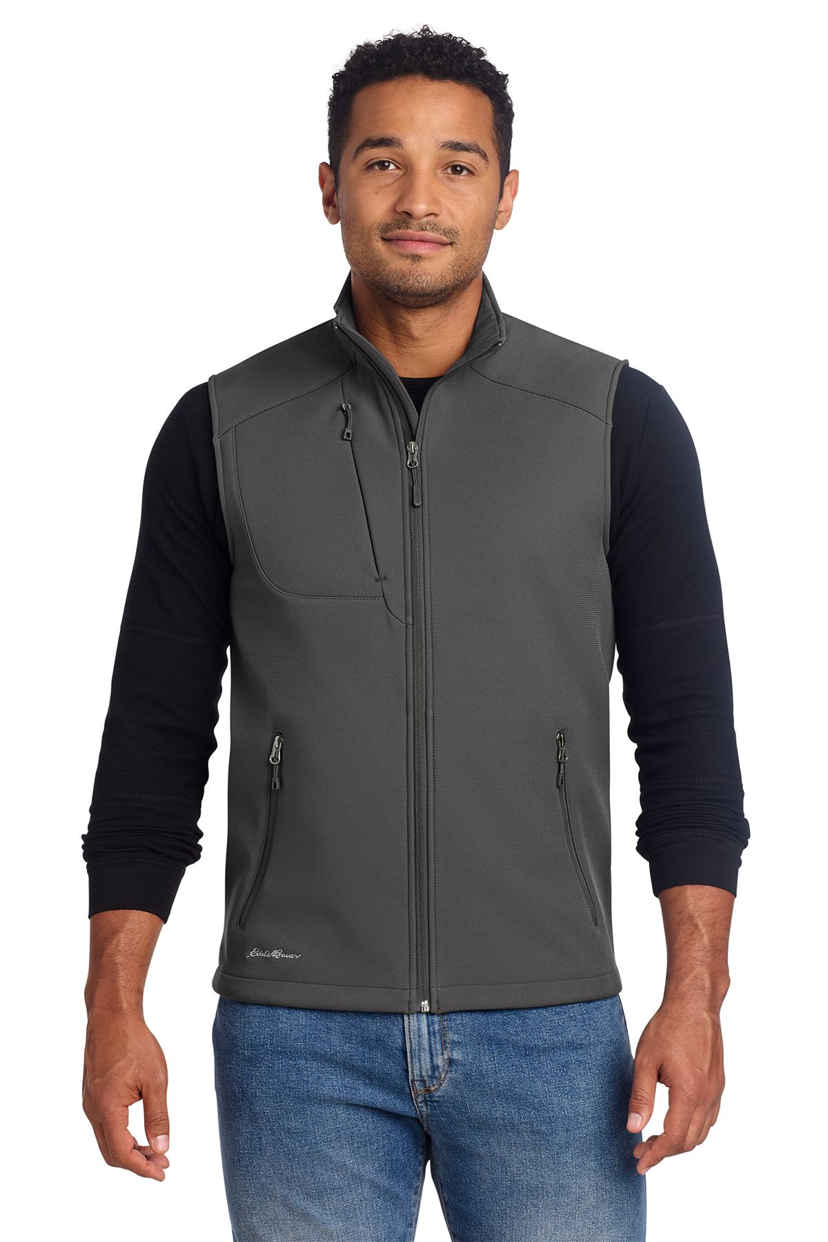 Man wearing a gray vest over a black long-sleeved shirt and jeans, smiling, standing against a white background.