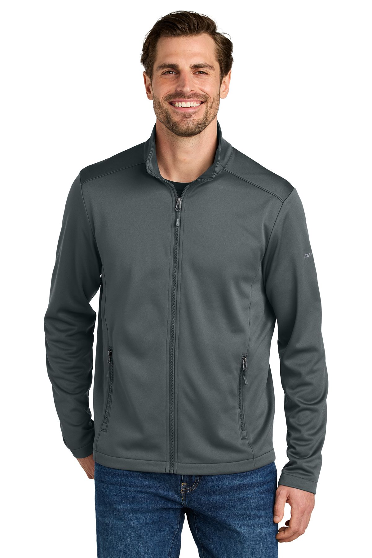Man in a dark blue zip-up jacket and jeans smiles, standing in front of a white background.
