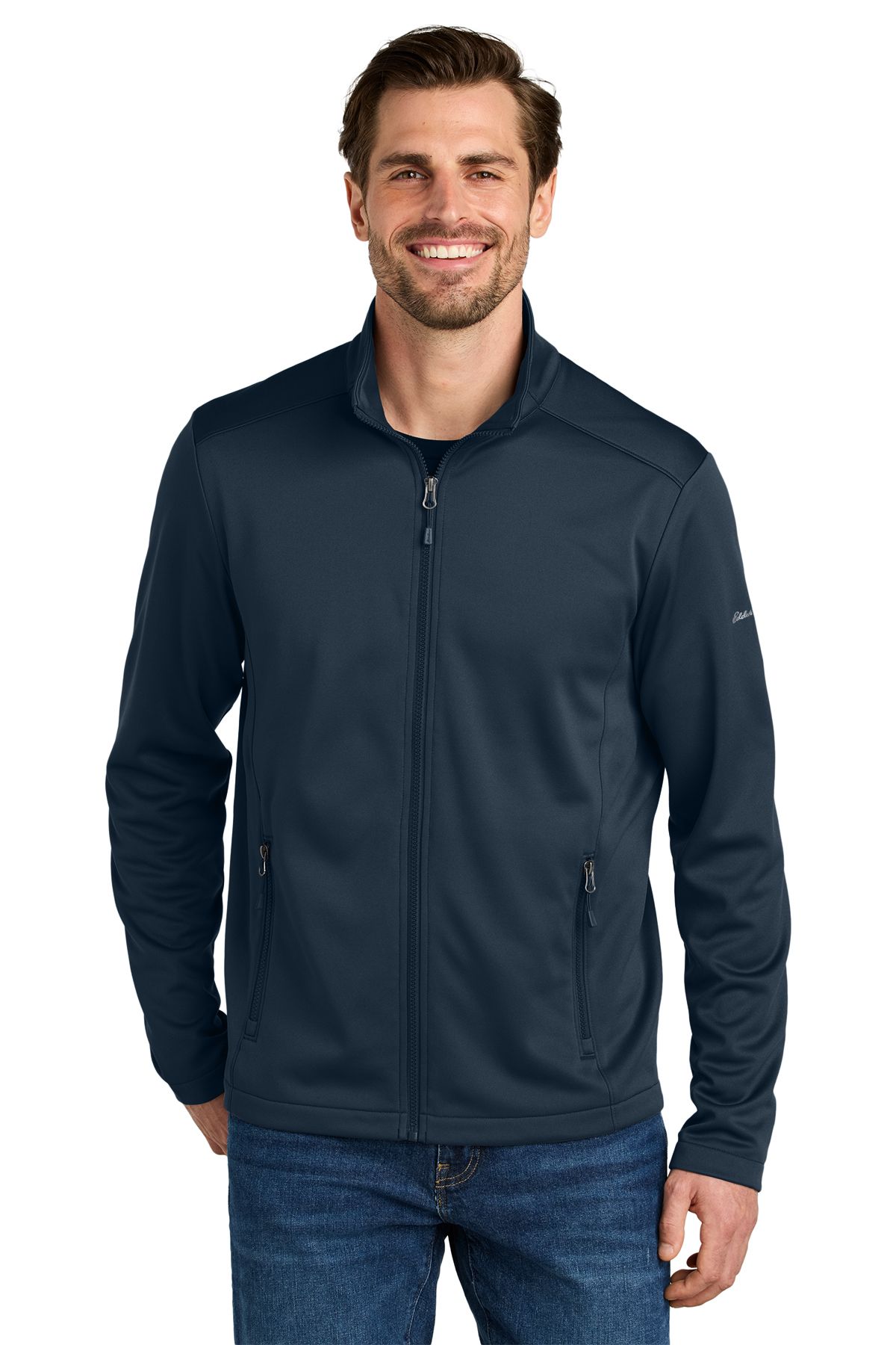 Man in a dark blue zip-up jacket and jeans smiles, standing in front of a white background.