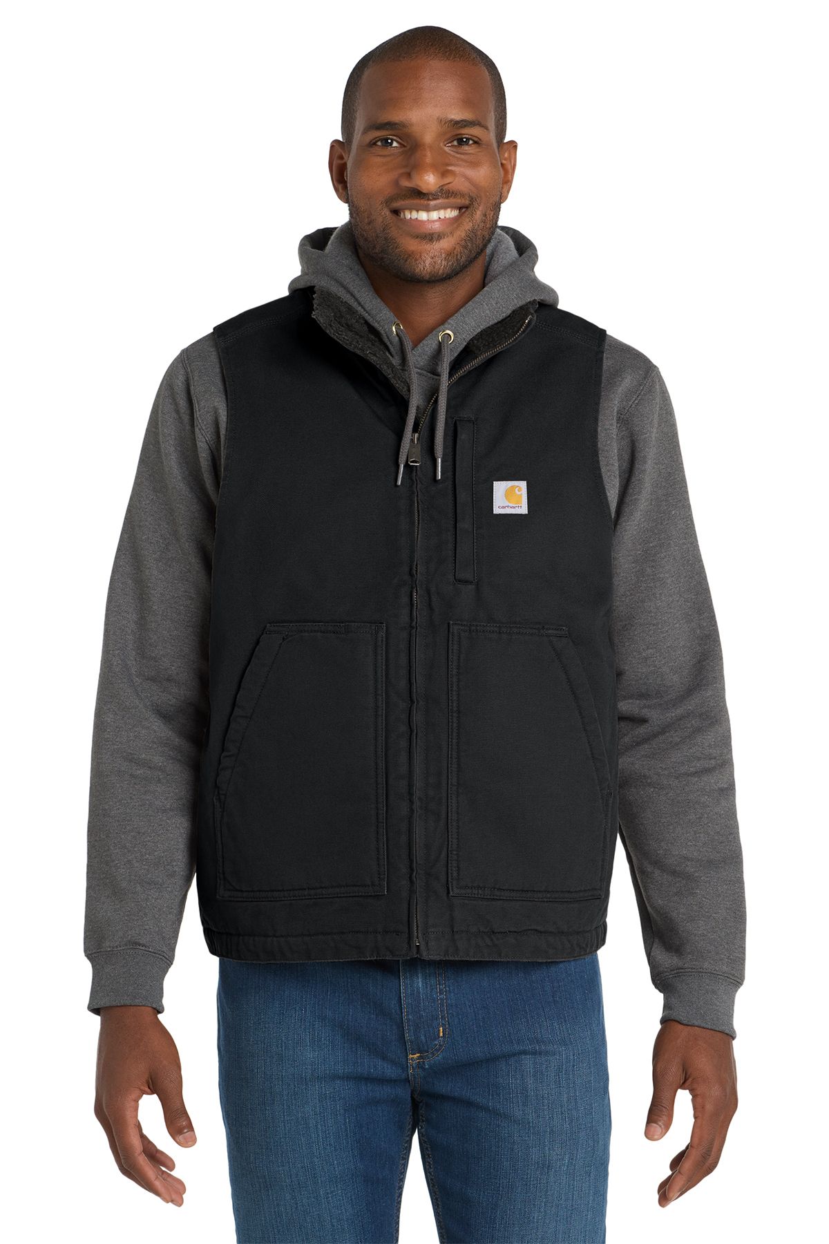 Man in brown Carhartt vest, gray hoodie, blue jeans. Smiling, indoors.