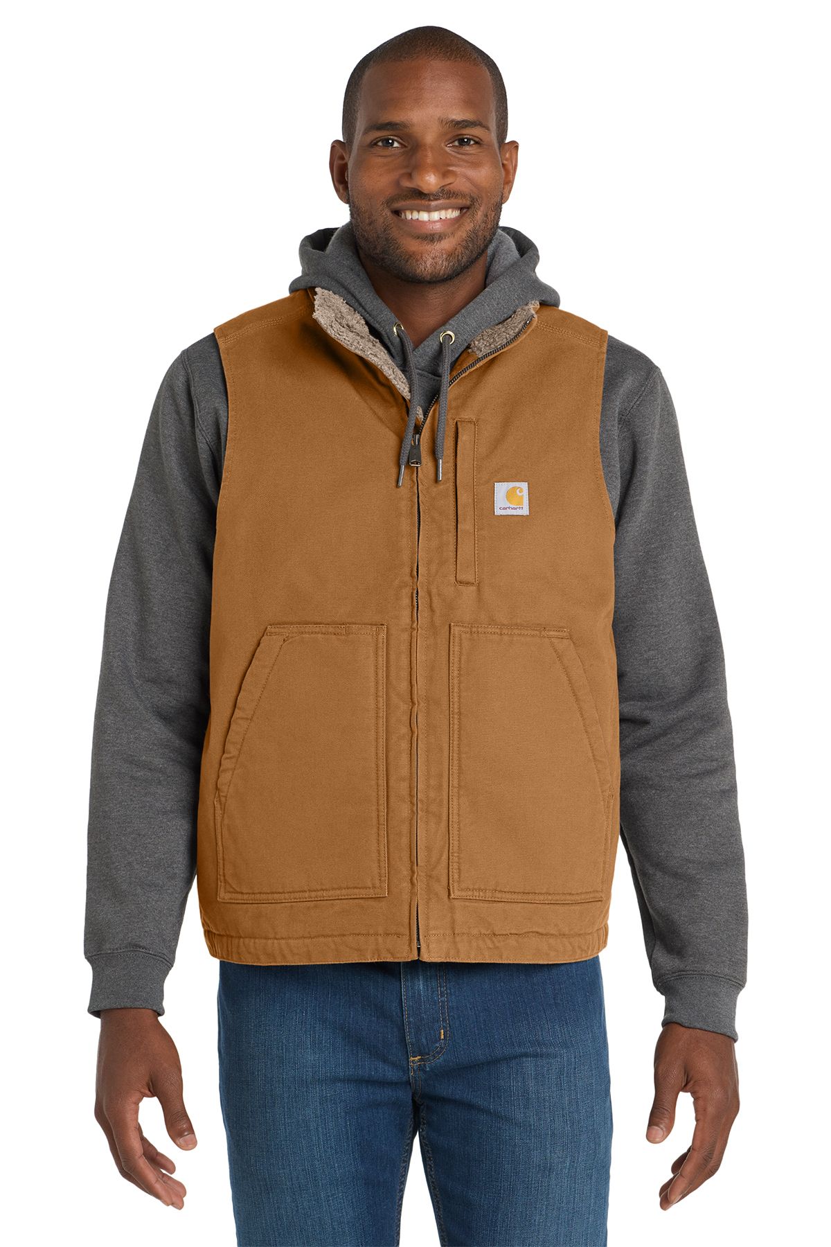 Man in brown Carhartt vest, gray hoodie, blue jeans. Smiling, indoors.