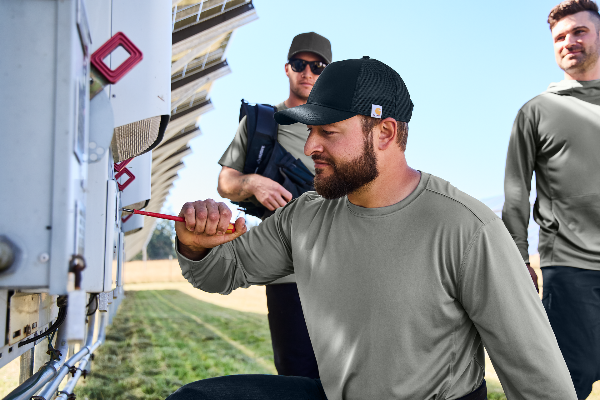 Man in cap working on solar panel with two others in field.