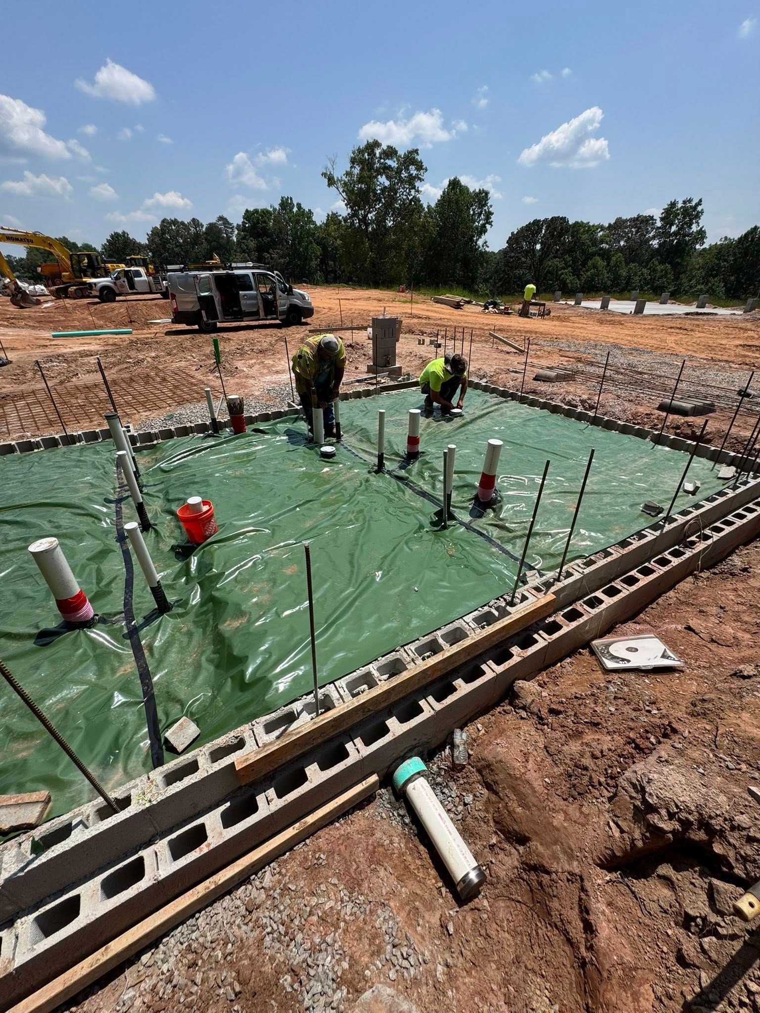 Workers in neon vests install plumbing pipes on a green-lined foundation site surrounded by blocks and dirt.