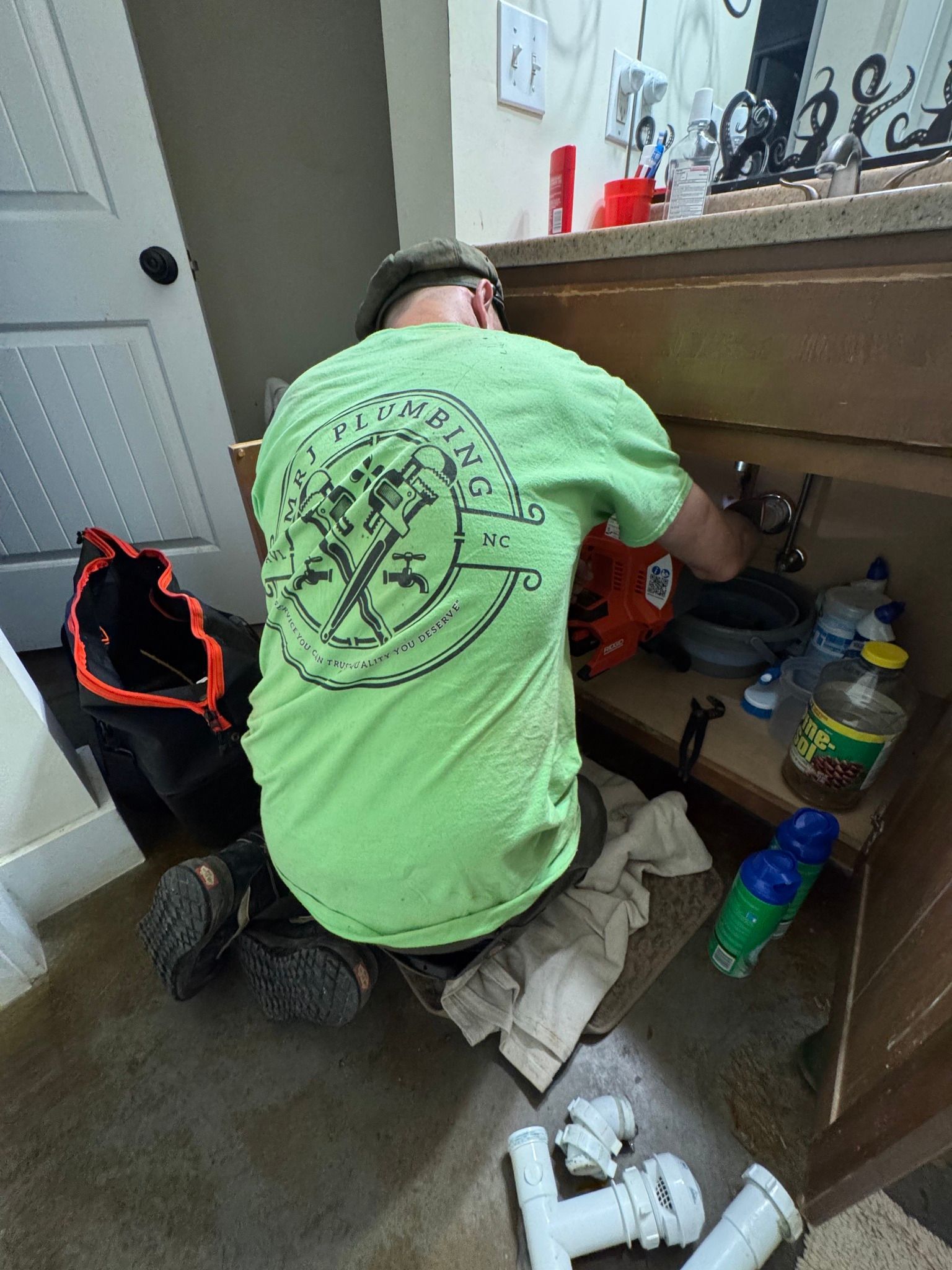 A person in a bright green t-shirt kneeling on the floor, fixing the plumbing underneath a bathroom sink.