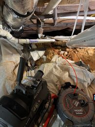Crawlspace plumbing repair in progress with a Ridgid K-5208 drum machine and Ridgid SeeSnake microReel camera staged next to an exposed cast-iron sewer line and PVC repair section.