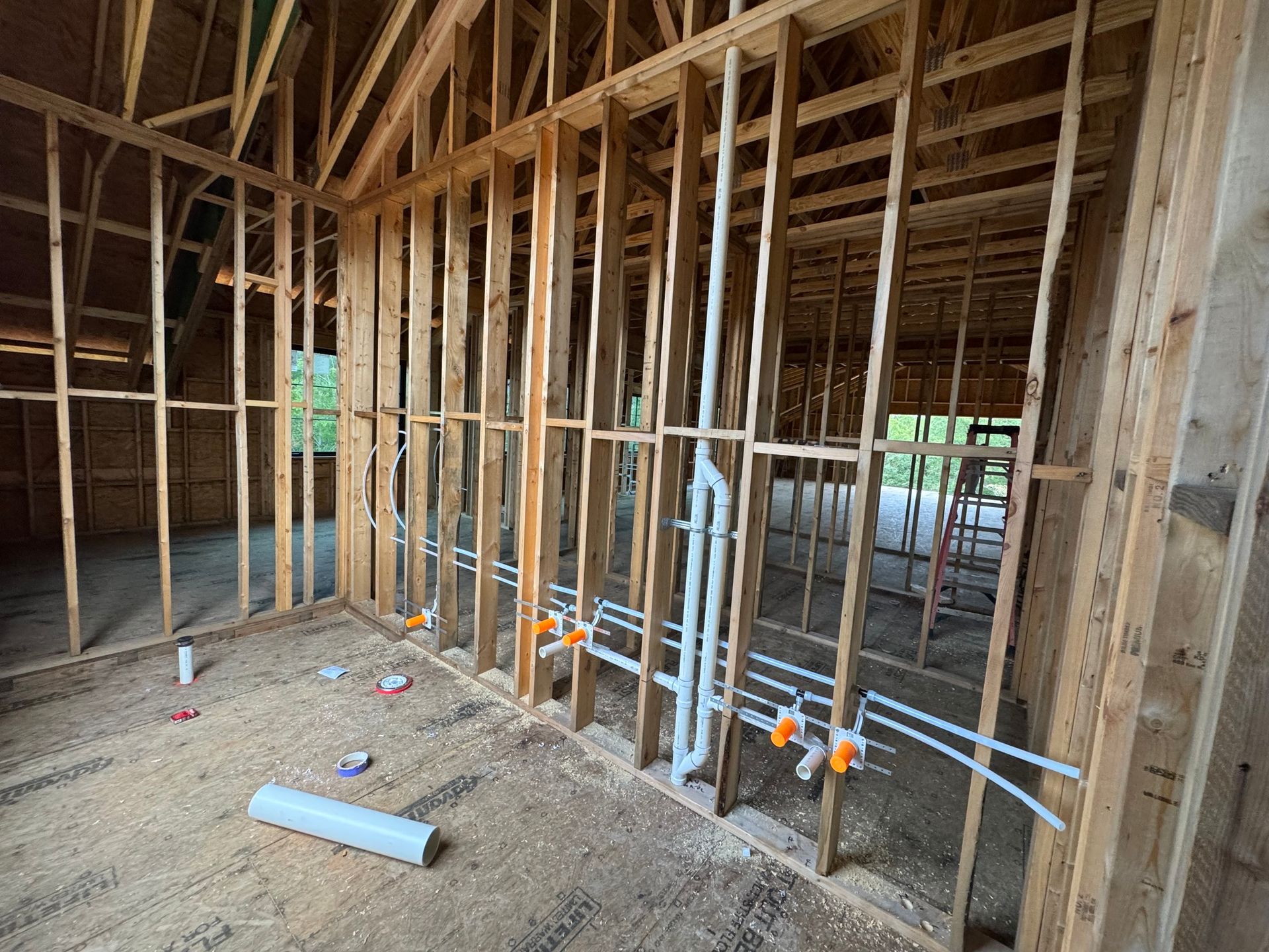 Unfinished wood-framed building interior showing exposed wall studs with installed plumbing pipes and drain lines.