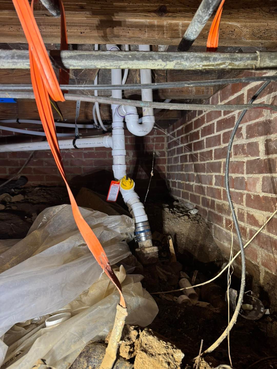 White PVC plumbing pipes with a yellow cleanout cap installed in a dirt-floor crawl space against a brick wall.