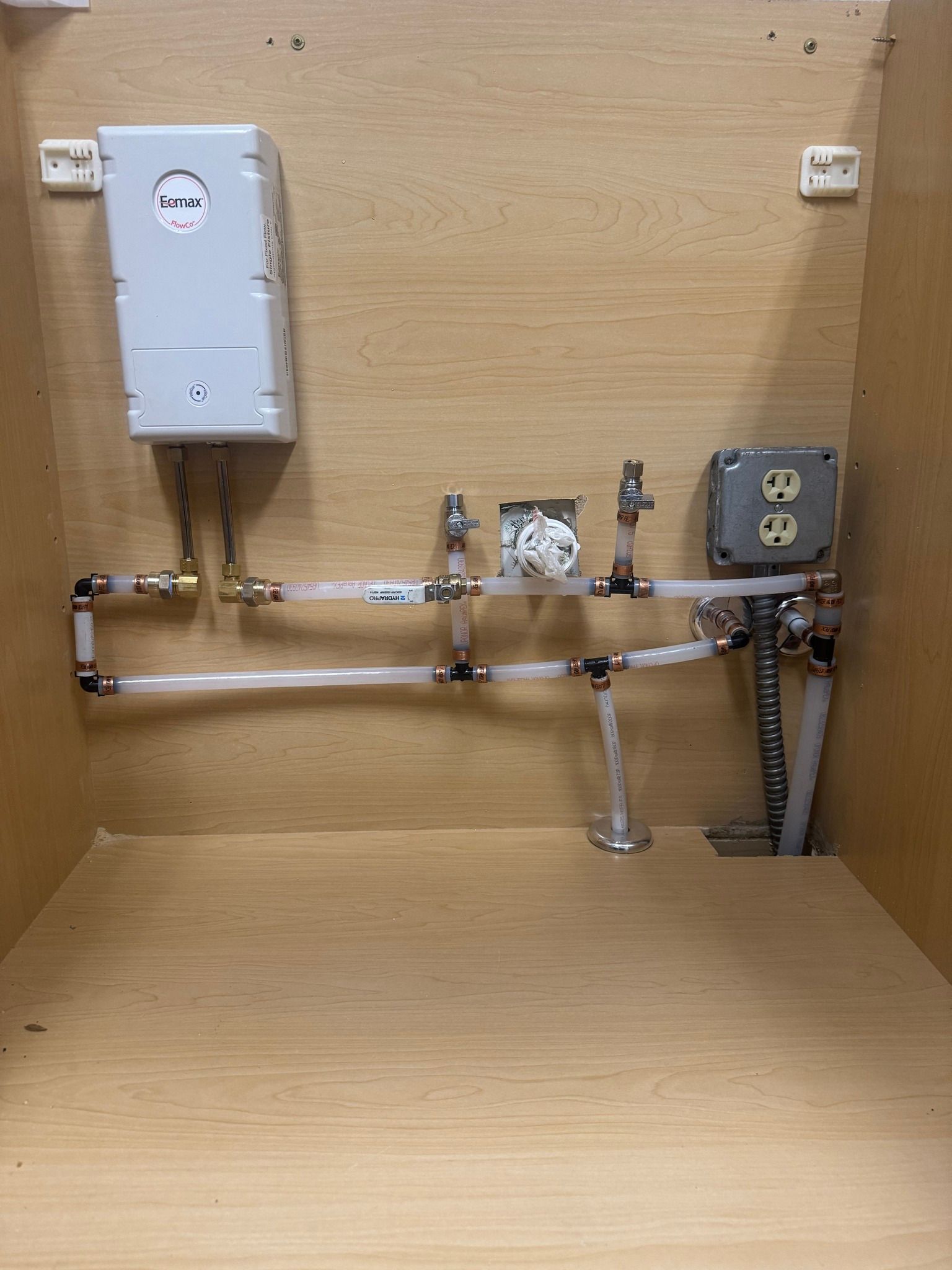Inside a wooden cabinet, a white electric tankless water heater is connected to white PEX pipes, valves, and a wall outlet.