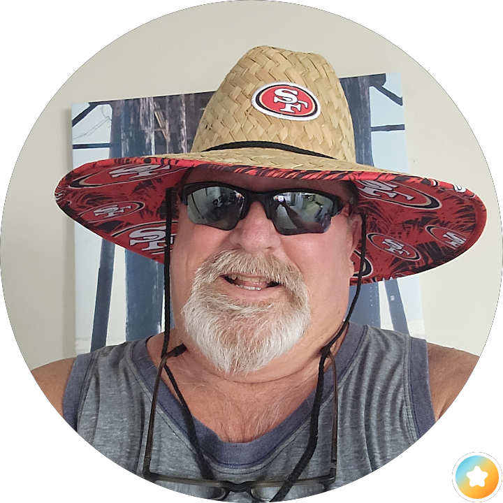 Man wearing a red San Francisco 49ers sun hat, black sunglasses, and a sleeveless gray shirt, smiling indoors