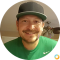 Smiling man in a black cap and green shirt indoors, with a ceiling fan in the background