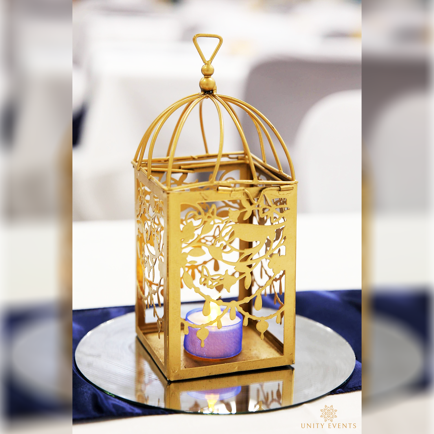 Gold birdcage-style lantern with a lit candle on a mirrored tray on a table