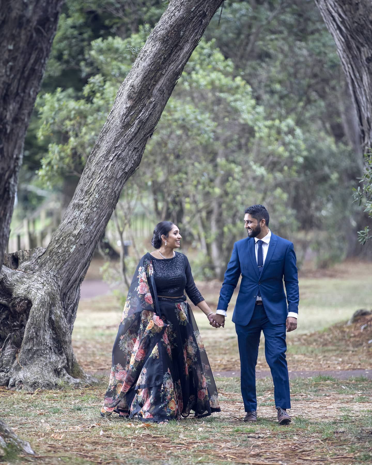 Couple holding hands in a wooded park, dressed in formal wear, looking at each other.