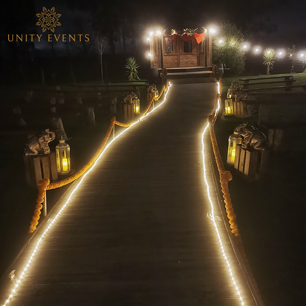 Lantern-lit wooden walkway leading to a warmly lit entrance at night, with string lights and greenery