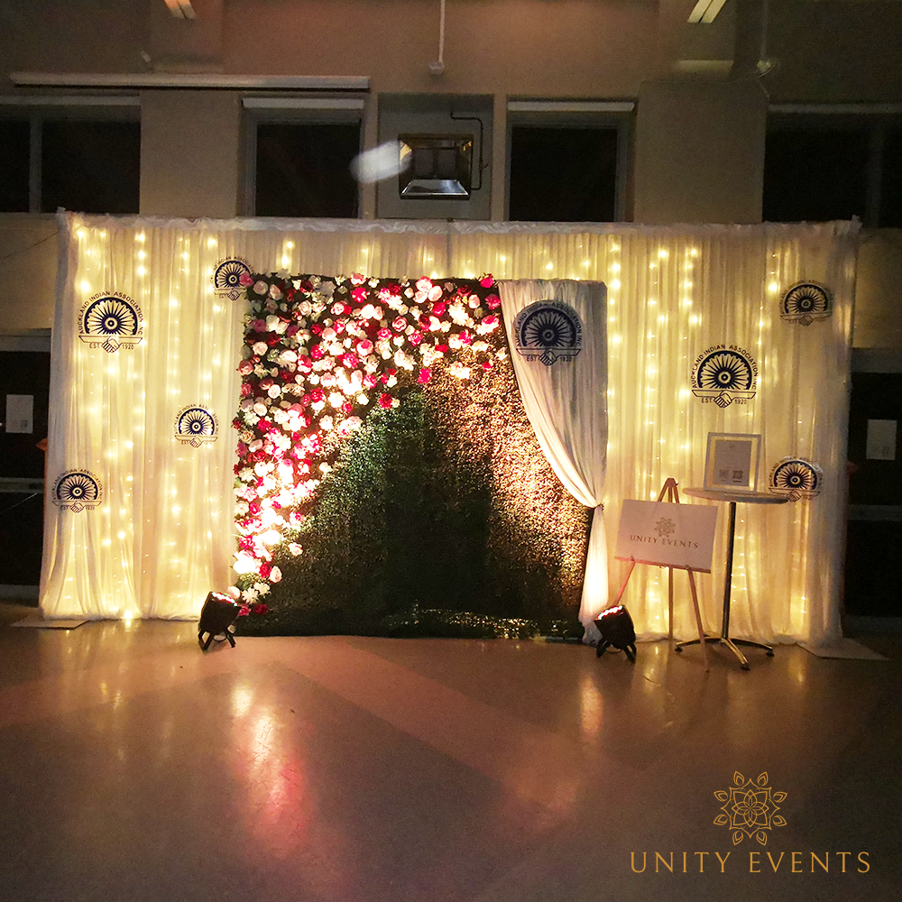 Elegant event photo backdrop with white drapes, warm lights, and red floral greenery centerpiece at night