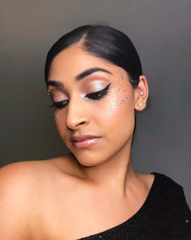 Close-up portrait of a woman with glitter makeup, eyes closed, wearing a black off-shoulder top against a gray background