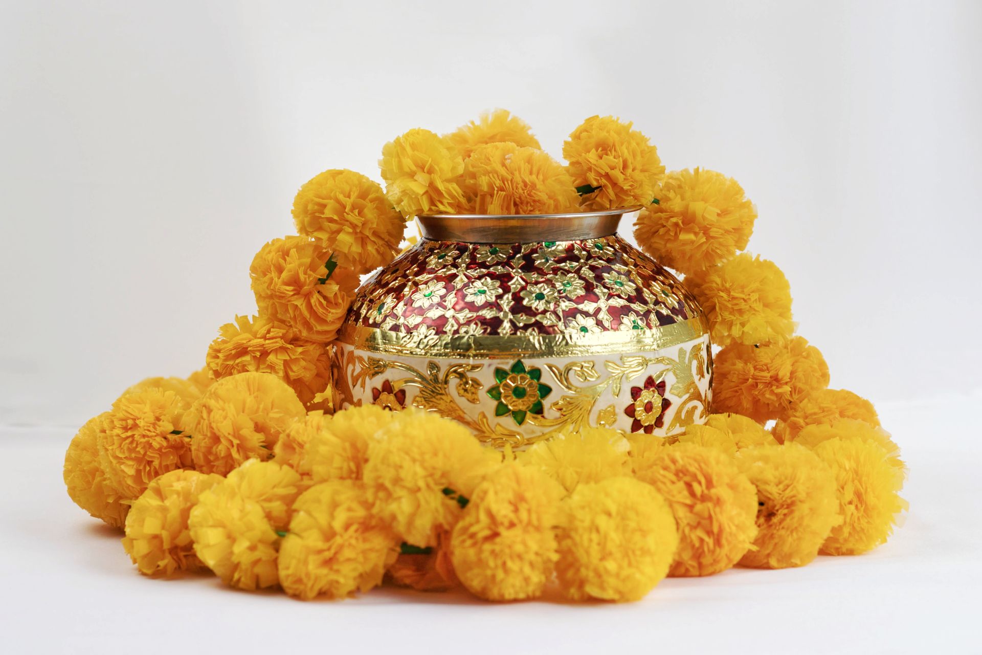 Decorated ceremonial pot surrounded by bright yellow marigold flowers on a white background