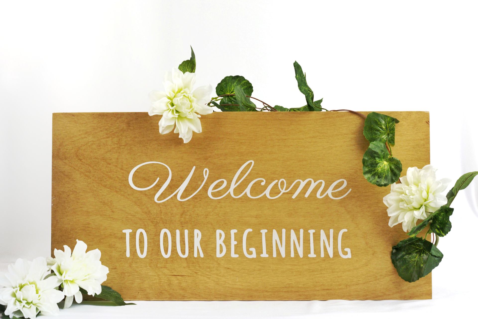 Wooden sign with white flowers and greenery reading “Welcome to our beginning”
